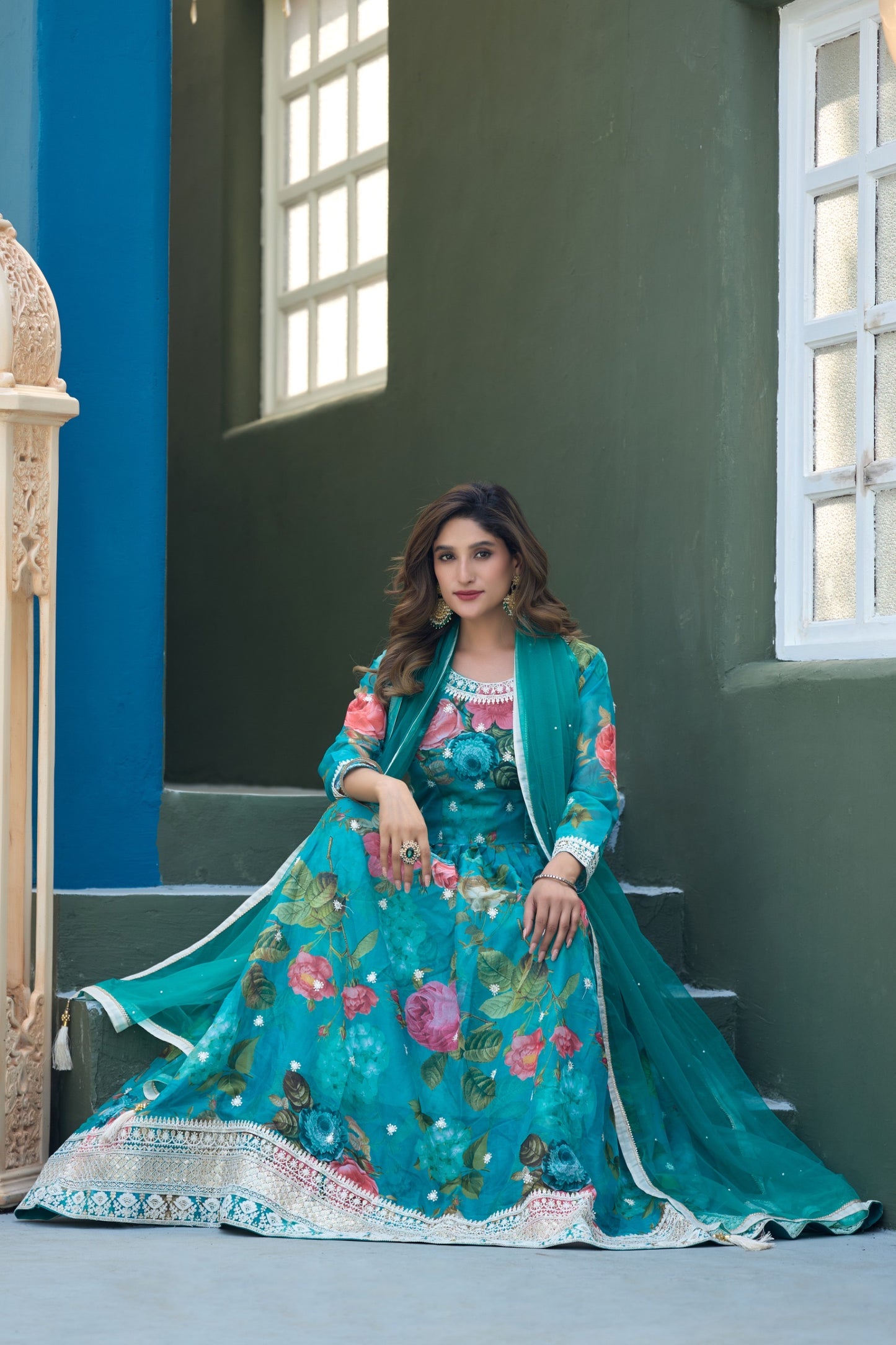 Woman in a teal floral dress sitting on steps with a blue wall and window in the background