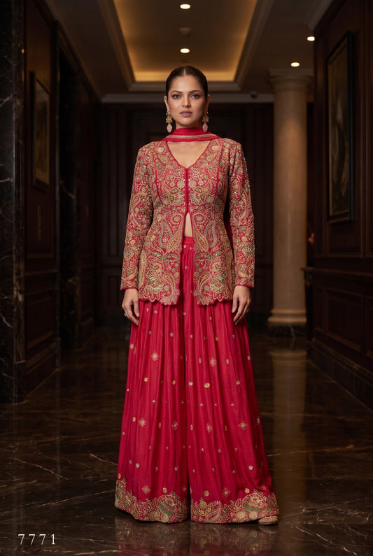A woman stands in a dark, elegant hallway wearing a traditional Indian outfit consisting of a red skirt with gold embroidery and a matching embroidered jacket, along with a red neck scarf.